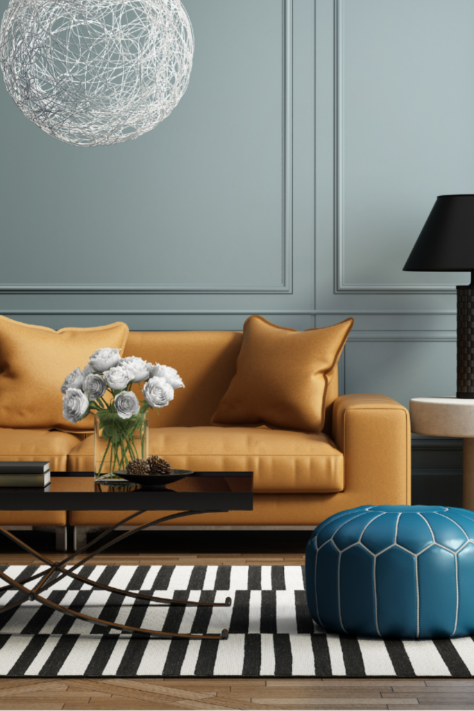 Eine pastellblaue Wand als Hintergrund, ein senfgelbes Sofa und eine schwarze Leselampe, modern und stilvoll eingerichtet. Im Vordergrund ein kleiner blauer Sitzhocker, der den minimalistischen Stil ergänzt.